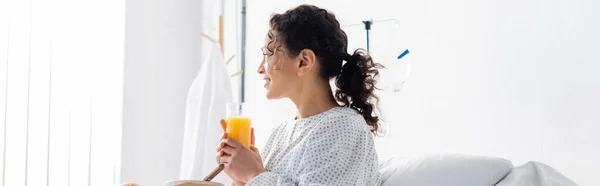 Smiling african american woman looking away while holding orange juice in hospital, banner — Stock Photo