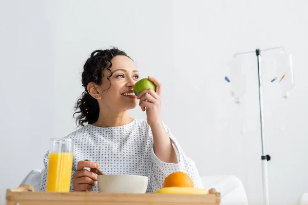 Alegre mulher afro-americana desfrutando de sabor de maçã fresca durante o café da manhã no hospital — Fotografia de Stock