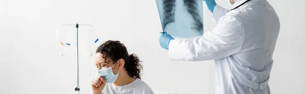 Doctor in latex gloves holding lungs x-ray near african american woman coughing in medical mask, banner — Stock Photo