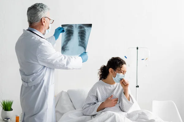 Radiologe mit medizinischer Maske hält Lungen-Röntgenbild in der Nähe afrikanisch-amerikanischer Maske hustet auf Krankenhausbett — Stockfoto