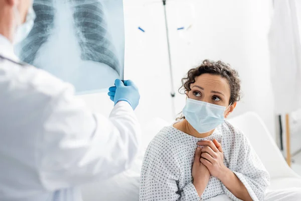 Besorgte Afroamerikanerin mit medizinischer Maske in der Nähe eines Radiologen in Latexhandschuhen, der die Lungen im verschwommenen Vordergrund röntgt — Stockfoto
