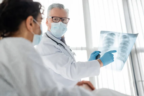 Radiologo in occhiali e maschera medica che punta ai polmoni radiografia vicino donna afroamericana in primo piano offuscata — Foto stock