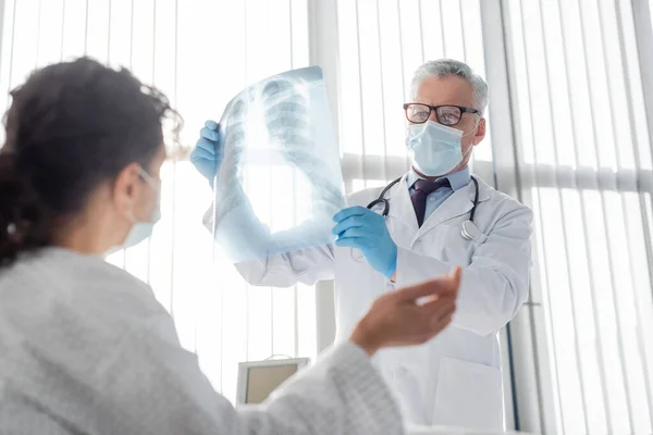 Radiólogo en máscara médica que sostiene los pulmones de rayos X cerca de la mujer afroamericana en primer plano borrosa - foto de stock