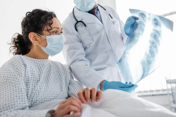 Radiólogo en guantes de látex mostrando rayos X de los pulmones a la mujer afroamericana en máscara médica, borrosa primer plano - foto de stock