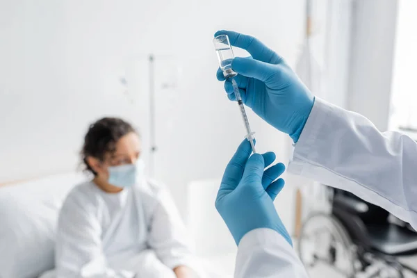 Mujer afroamericana borrosa con máscara médica cerca del médico con guantes de látex sosteniendo jeringa y frasco con vacuna - foto de stock