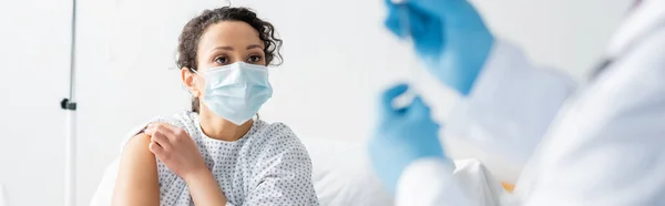 Mujer afroamericana en máscara médica cerca del médico en guantes de látex en primer plano borroso, bandera - foto de stock