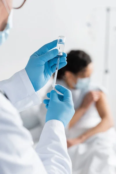Médico en guantes de látex que sostiene la jeringa y el frasco con la vacuna cerca borrosa mujer afroamericana en máscara médica - foto de stock