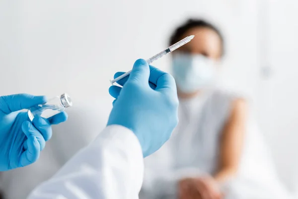 Femme afro-américaine floue près du médecin en gants de latex tenant bouteille avec vaccin et seringue — Photo de stock