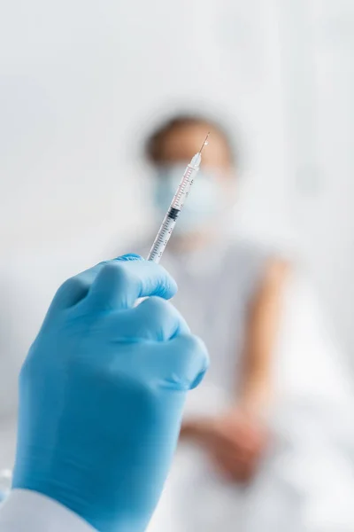 Jeringa con vacuna en la mano del médico en guante de látex cerca de la mujer afroamericana sobre fondo borroso - foto de stock