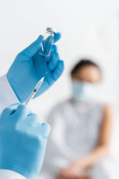 Medico in guanti di lattice in possesso di siringa e flacone con vaccino vicino alla donna afroamericana su sfondo sfocato — Foto stock