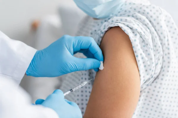 Частичный взгляд врача в латексных перчатках, делающего инъекцию вакцины женщине в медицинской маске — стоковое фото