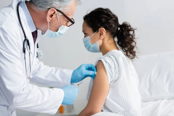 Médico maduro em máscara médica fazendo injeção de vacina para mulher afro-americana — Fotografia de Stock
