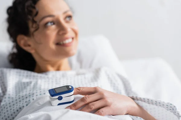 Donna afro-americana sorridente con pulsossimetro al dito distogliendo lo sguardo in ospedale, sfondo sfocato — Foto stock