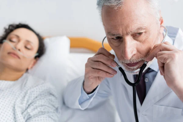 Doctor poniendo en estetoscopio cerca enfermo africano americano mujer en borrosa fondo - foto de stock