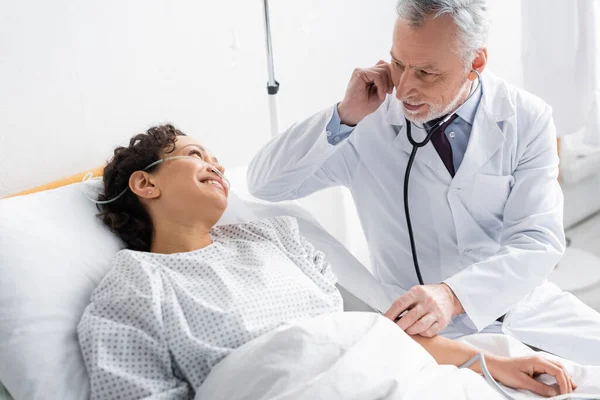 Arzt untersucht Afroamerikanerin lächelnd im Krankenhausbett — Stockfoto