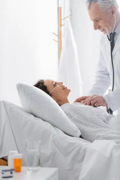 Sonriente doctor examinando positivo africano americano mujer con estetoscopio en clínica - foto de stock