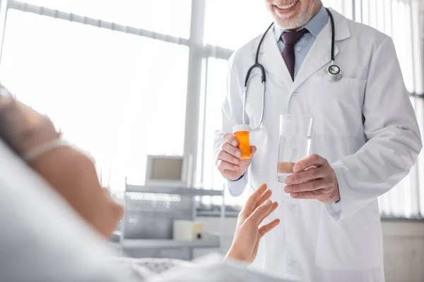 Vue recadrée d'un médecin souriant tenant des pilules et un verre d'eau près d'une femme afro-américaine floue — Photo de stock