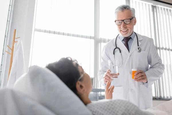 Médico maduro sosteniendo agua y medicamentos cerca de la mujer afroamericana en primer plano borroso - foto de stock