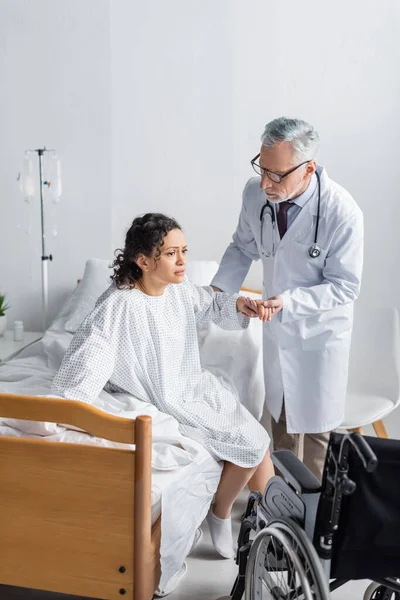 Médico ayudando a una mujer afroamericana a levantarse de la cama cerca de una silla de ruedas en el hospital - foto de stock