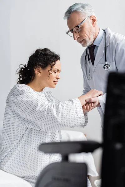 Attentive doctor supporting african american woman frowning from pain near wheelchair on blurred foreground — Stock Photo