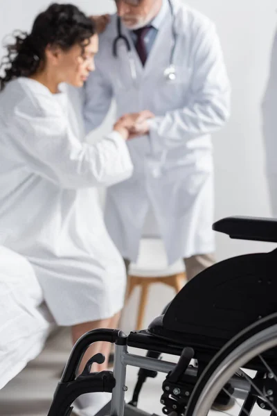 Foco seletivo da cadeira de rodas perto do médico ajudando a mulher afro-americana se levantar da cama na clínica — Fotografia de Stock