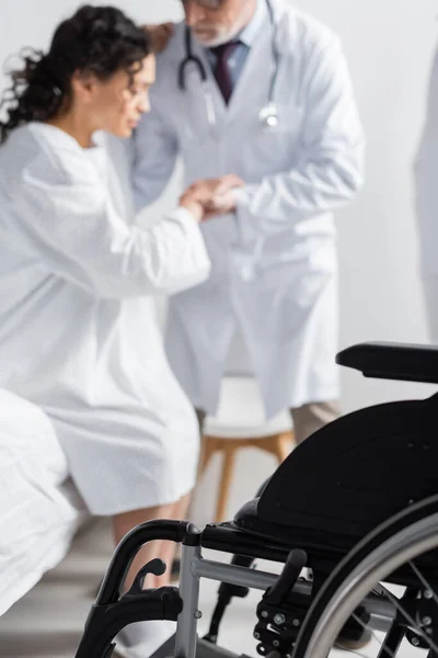 Selective focus of wheelchair near doctor helping african american woman getting up from bed in clinic — Stock Photo