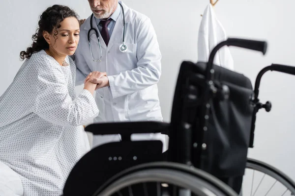 Médecin soutenant femme afro-américaine près de fauteuil roulant sur le premier plan flou — Photo de stock