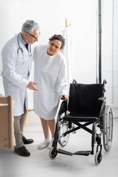 Doctor pointing at wheelchair while supporting african american woman in hospital — Stock Photo