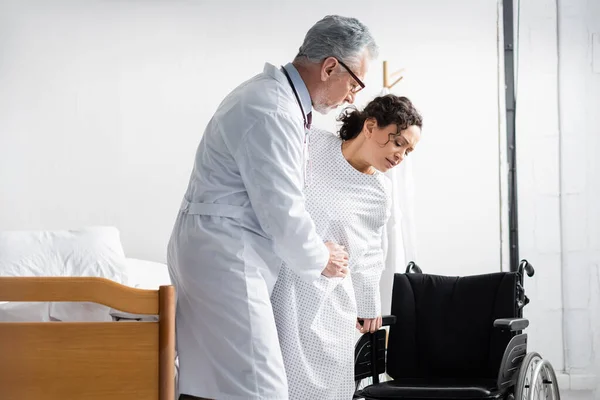 Médecin mature soutenant femme afro-américaine près de fauteuil roulant en clinique — Photo de stock