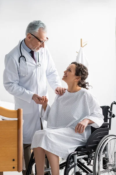 Médico maduro cogido de la mano de la sonriente mujer afroamericana sentada en silla de ruedas - foto de stock