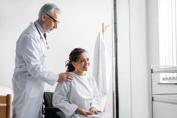 Medico sorridente toccare spalla di felice donna africana americana seduto in sedia a rotelle — Foto stock