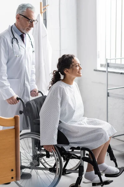 Sonriente doctor cerca feliz africano americano mujer sentado en silla de ruedas en hospital - foto de stock