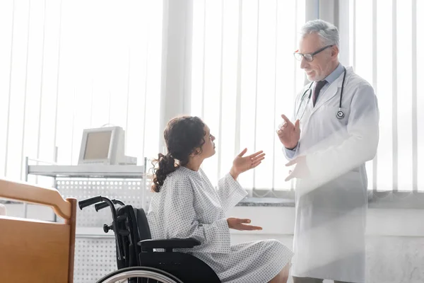 Mujer afroamericana en silla de ruedas gesticulando mientras habla con el médico en el hospital - foto de stock
