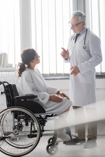 Mature doctor talking to african american woman sitting in wheelchair on blurred foreground — Stock Photo