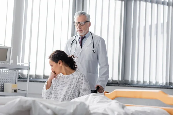 Verärgerte Afroamerikanerin sitzt im Rollstuhl in der Nähe von Arzt und Krankenhausbett auf verschwommenem Vordergrund — Stockfoto