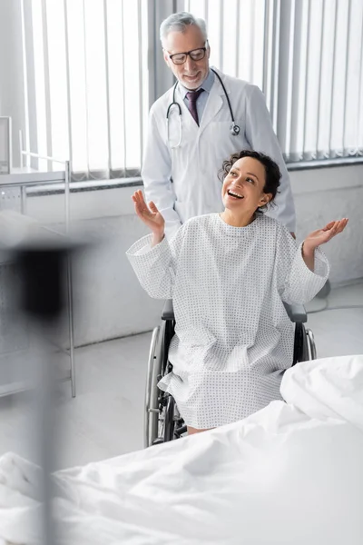 Femme afro-américaine gaie en fauteuil roulant montrant geste wow près du médecin, premier plan flou — Photo de stock