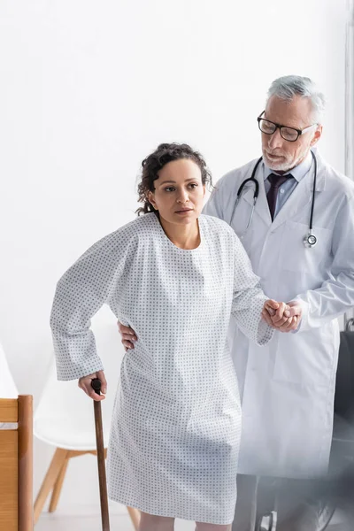 Doctor helping african american doctor walking with stick in hospital — Stock Photo