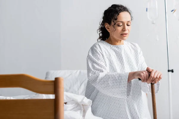 African american woman with walking stick sitting on bed in hospital, blurred foreground — Stock Photo
