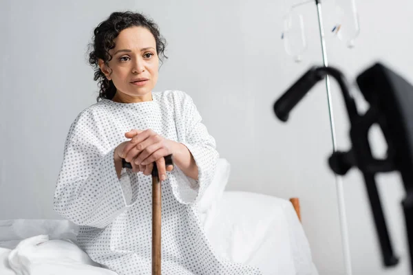 Upset african american woman sitting on hospital bed with walking stick on blurred foreground — Stock Photo