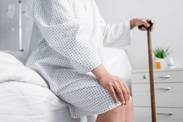 Vista recortada de la mujer afroamericana cogida de la mano sobre la rodilla mientras está sentada en la cama con bastón - foto de stock