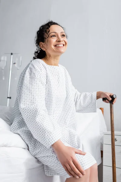 Joyful african american woman touching knee while sitting on bed with walking stick — Stock Photo