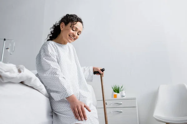 Positivo afroamericano donna toccare ginocchio mentre seduto sul letto con bastone da passeggio — Foto stock