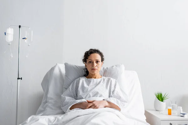 Kranke Afroamerikanerin blickt in Kamera, während sie im Krankenhausbett in der Nähe des Drop Counter sitzt — Stockfoto