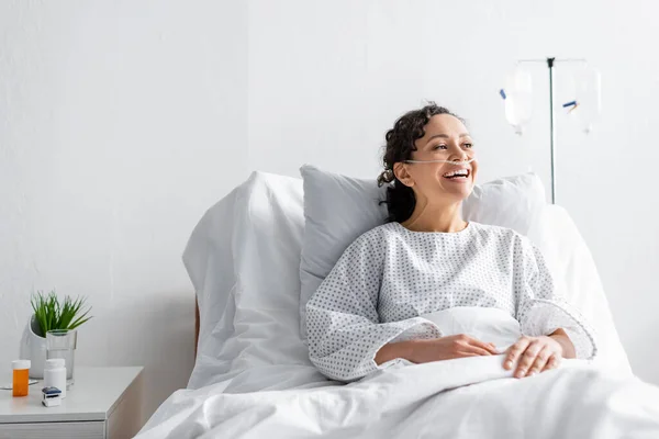 Feliz africano americano mujer riendo mientras sentado en hospital cama - foto de stock