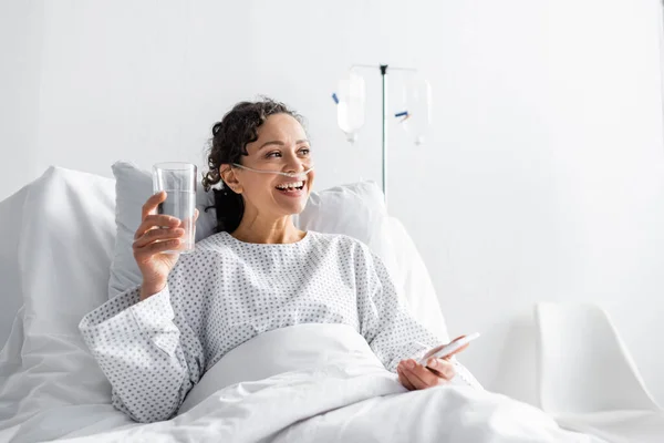 Felice donna afroamericana sorridente mentre tiene in mano un bicchiere d'acqua e un telefono cellulare in clinica — Foto stock
