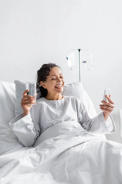 Freudige afrikanisch-amerikanische Frau blickt auf Smartphone, während sie im Krankenhaus ein Glas Wasser in der Hand hält — Stockfoto