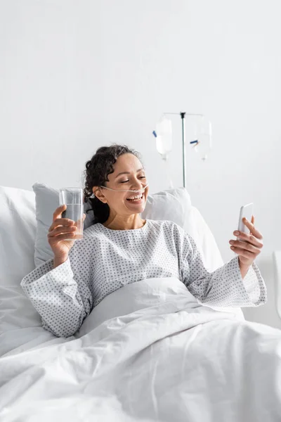 Alegre mujer afroamericana mirando el teléfono inteligente mientras sostiene un vaso de agua en el hospital - foto de stock