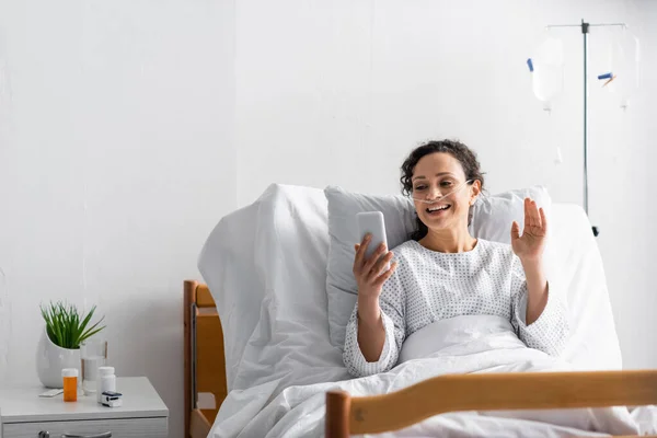 Sonriente mujer afroamericana agitando la mano durante la videollamada en el teléfono inteligente en el hospital - foto de stock