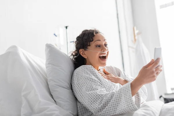 Amazed african american woman touching chest while holding smartphone in clinic — Stock Photo