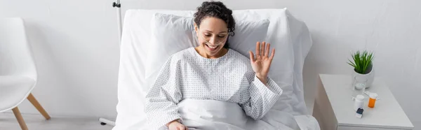 Femme afro-américaine joyeux agitant la main tout en étant assis dans le lit d'hôpital, bannière — Photo de stock